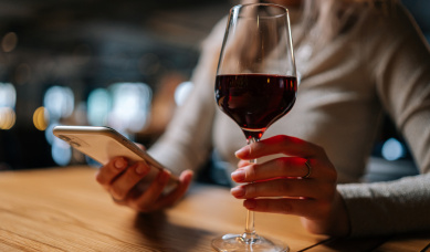 Close Up Cropped Shot Of Unrecognizable Young Woman Using Smartphone, Typing Online Message Sitting At Table Holding In Hand Glass Of Red Wine At Restaurant.