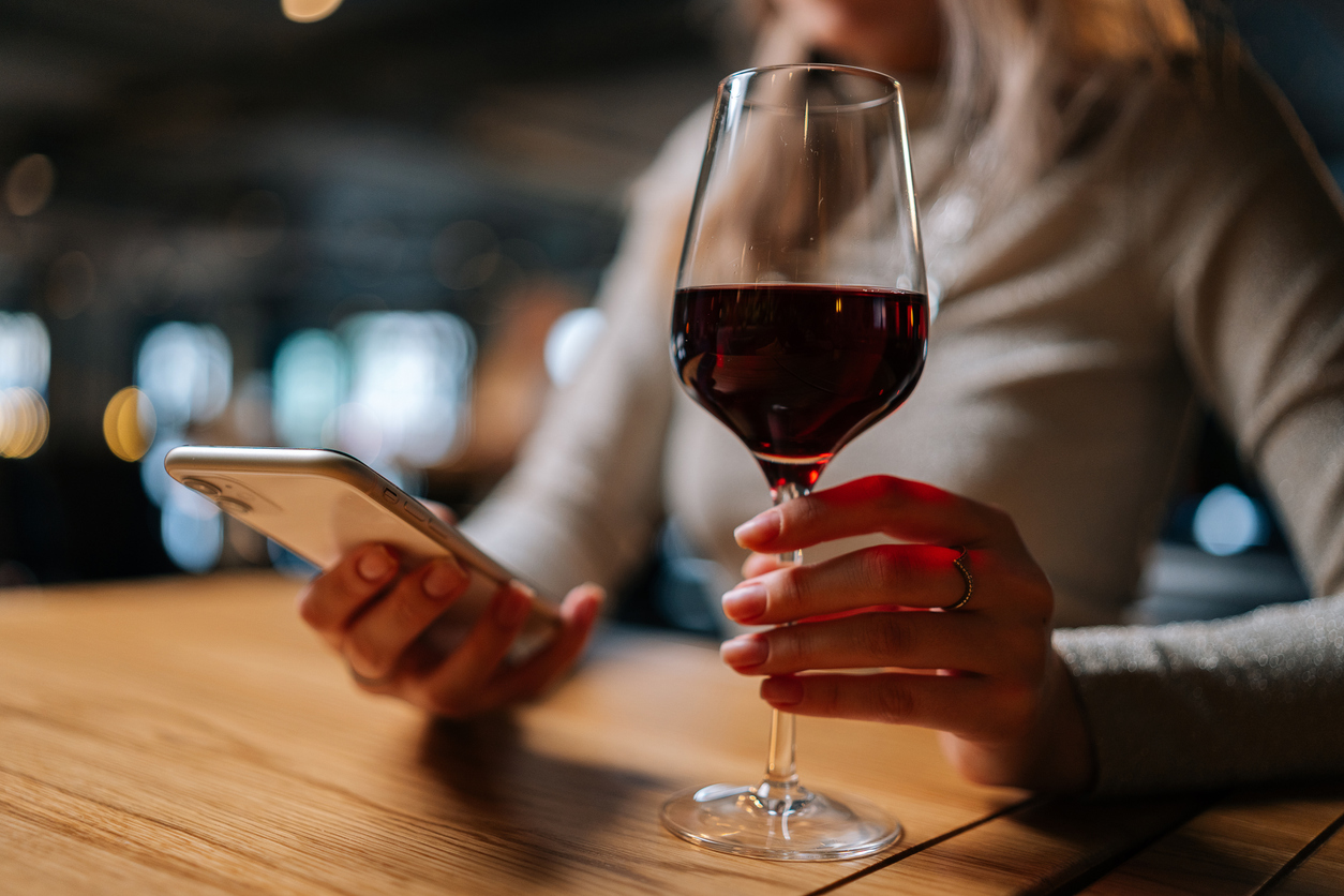 Close Up Cropped Shot Of Unrecognizable Young Woman Using Smartphone, Typing Online Message Sitting At Table Holding In Hand Glass Of Red Wine At Restaurant.